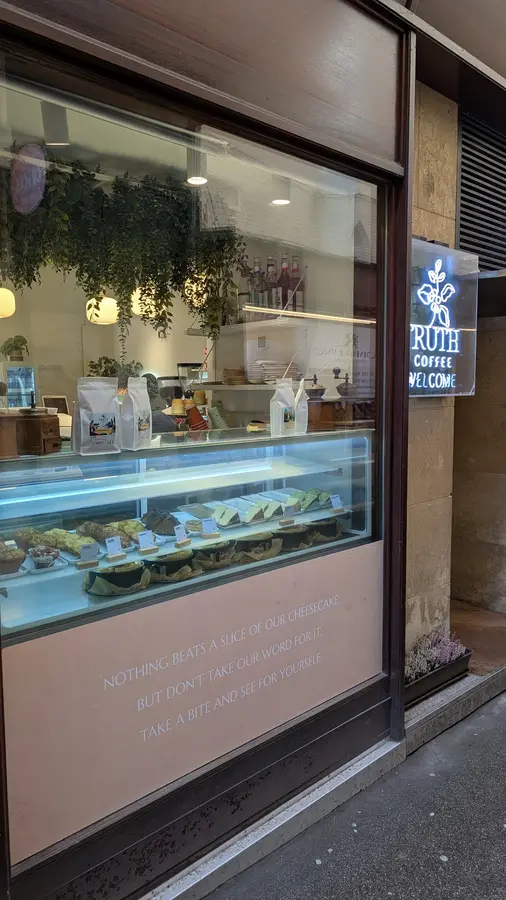 Schaufenster mit Kuchenvitrine, Leuchtschild „Truth Coffee Welcome“ und dem Cheesecake-Spruch